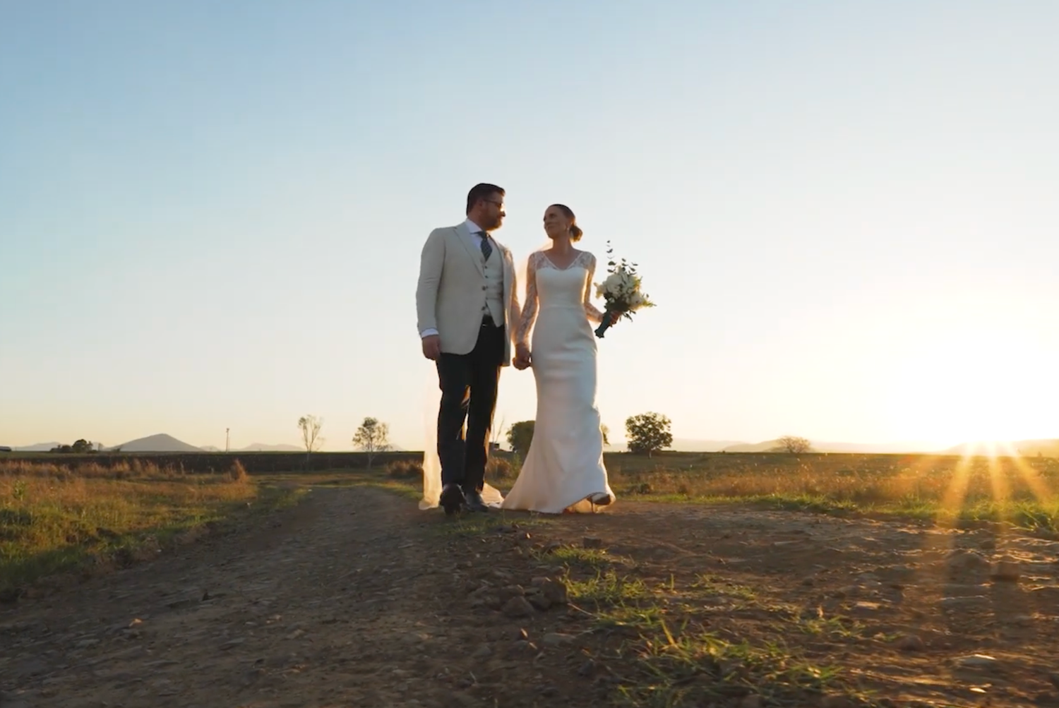 A couple in wedding attire walks down a dirt path at sunset holding hands while the bride carries a bouquet.
