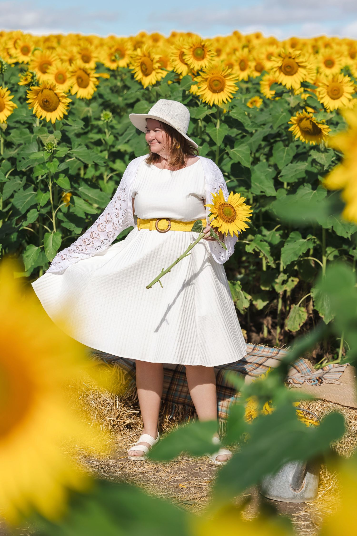 Woman in white dress and hat holding a sunflower in a vibrant sunflower field on a sunny day.