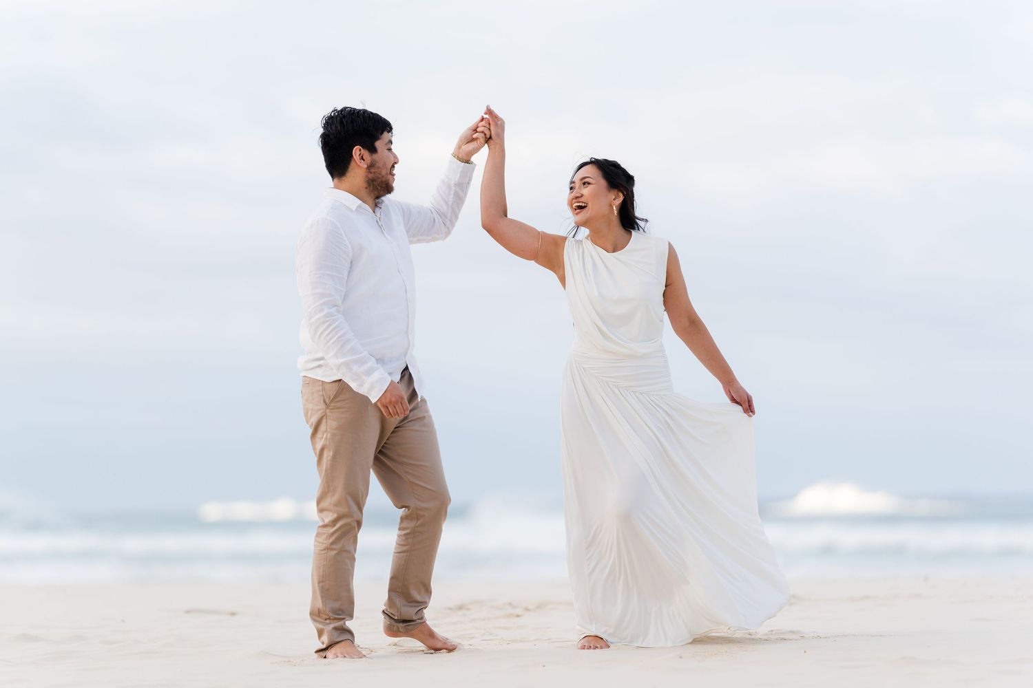 Couple dancing on beach in white outfits, holding hands raised, waves in background, romantic wedding photo.