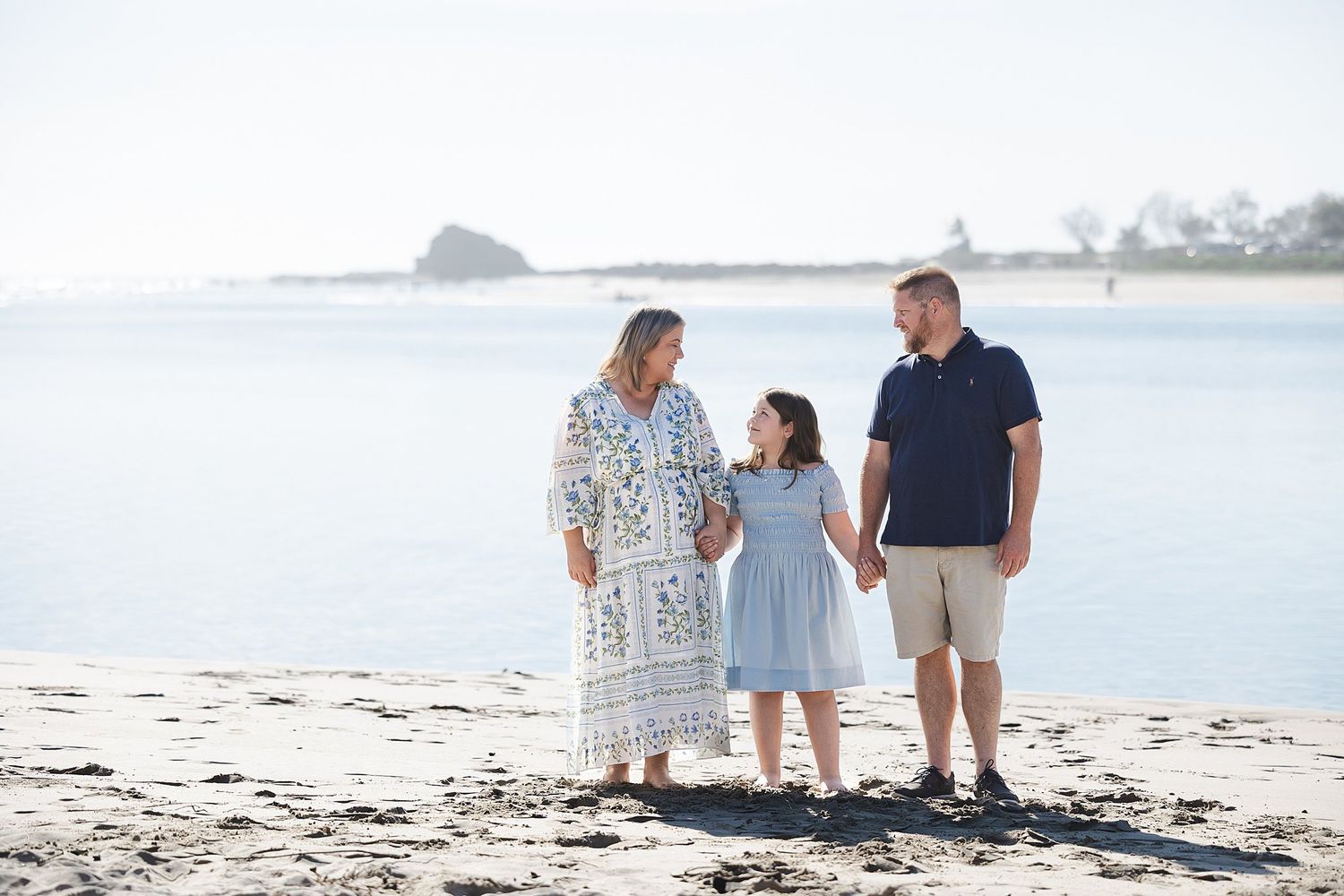 A family walks together on a sunny beach with calm waters and distant shoreline in the background.