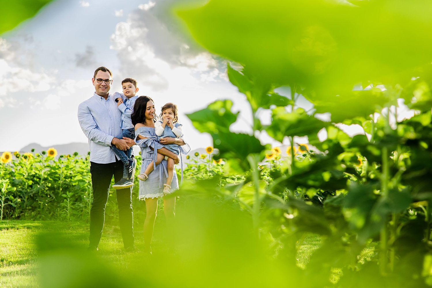 A family stands together in a field framed by lush green foliage with dramatic clouds in the background.