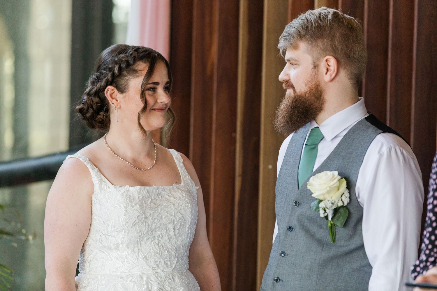 A bride in a white lace gown and groom in a gray vest with green tie share a loving glance at their wedding.