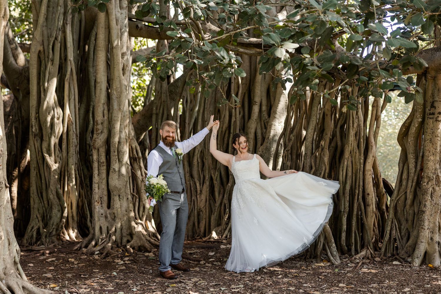Bride and groom hold hands dancing under sprawling banyan tree roots during outdoor wedding celebration.