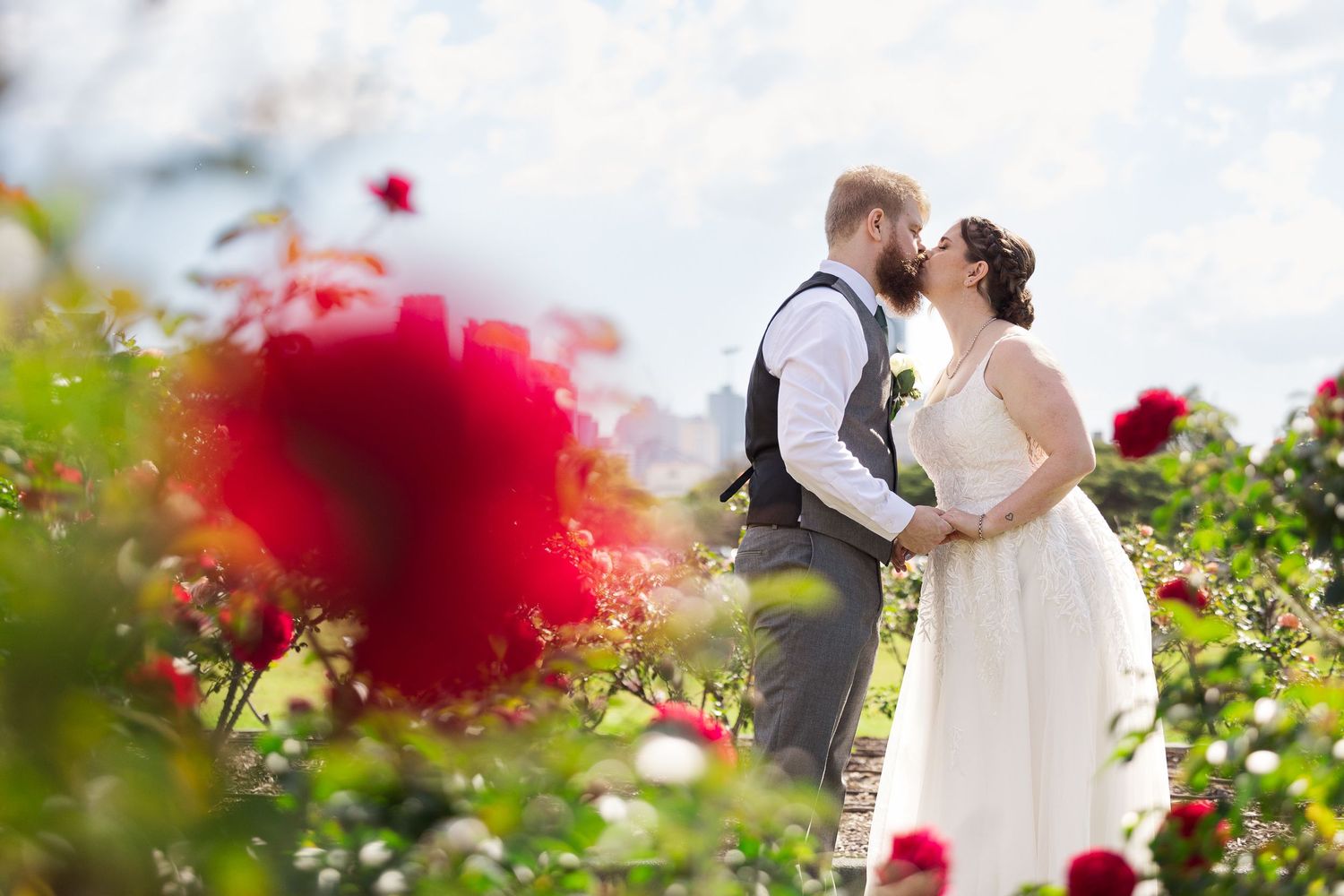Bride and groom kissing in a lush rose garden with vibrant red blooms framing the romantic outdoor wedding moment.