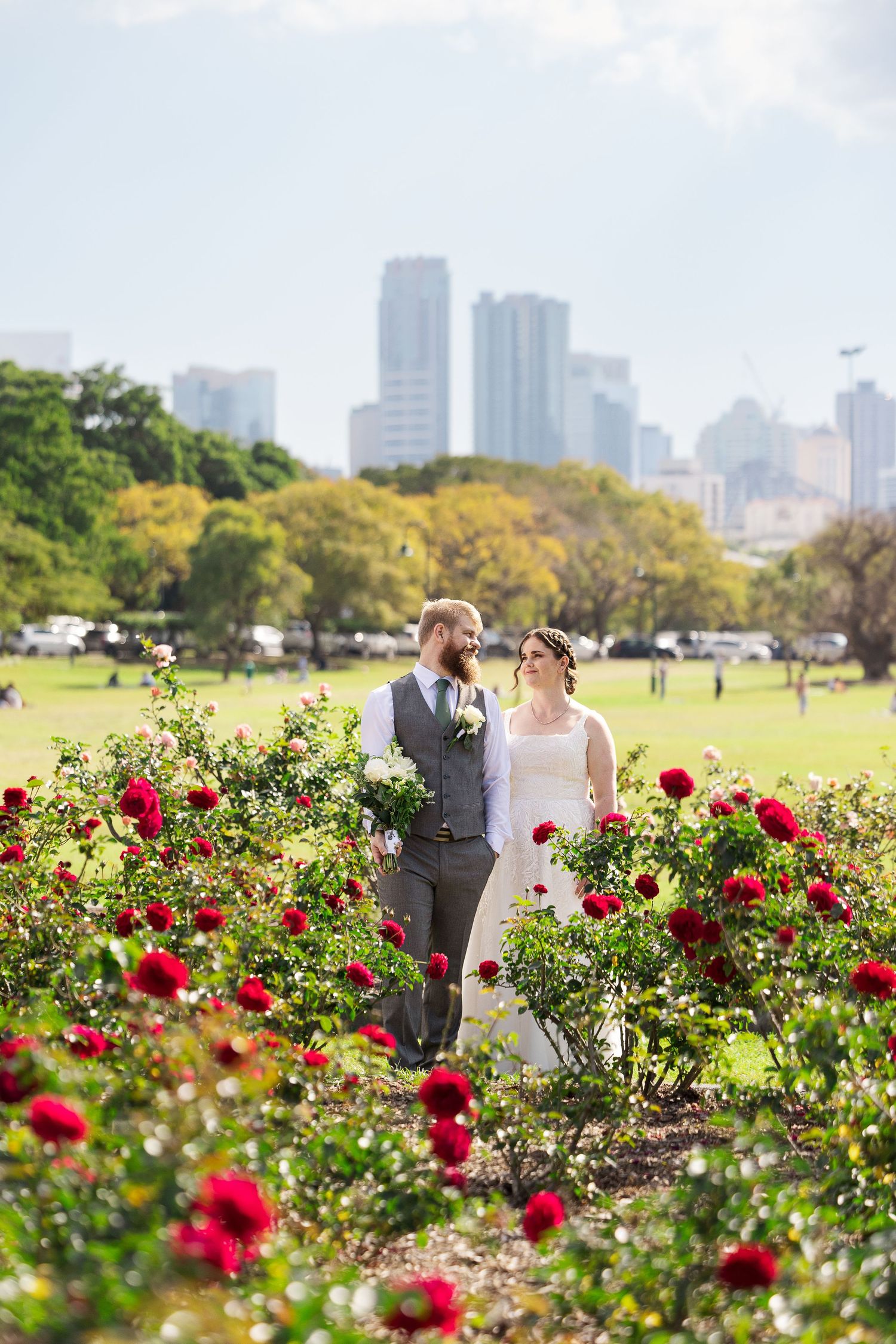 Bride and groom standing among red roses in a park with city skyline backdrop on their wedding day.