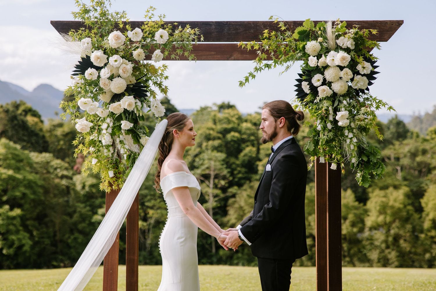 A couple exchanges vows during an outdoor wedding ceremony beneath a floral arch with mountain views in the background.