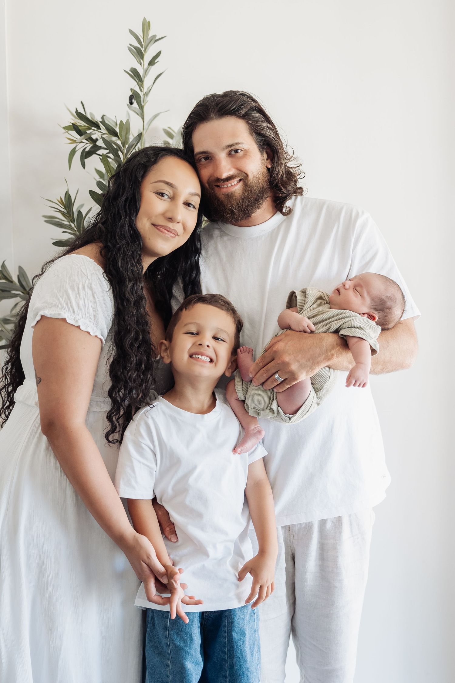 A family of four wearing white shirts poses together against a white wall with olive branches in the background.
