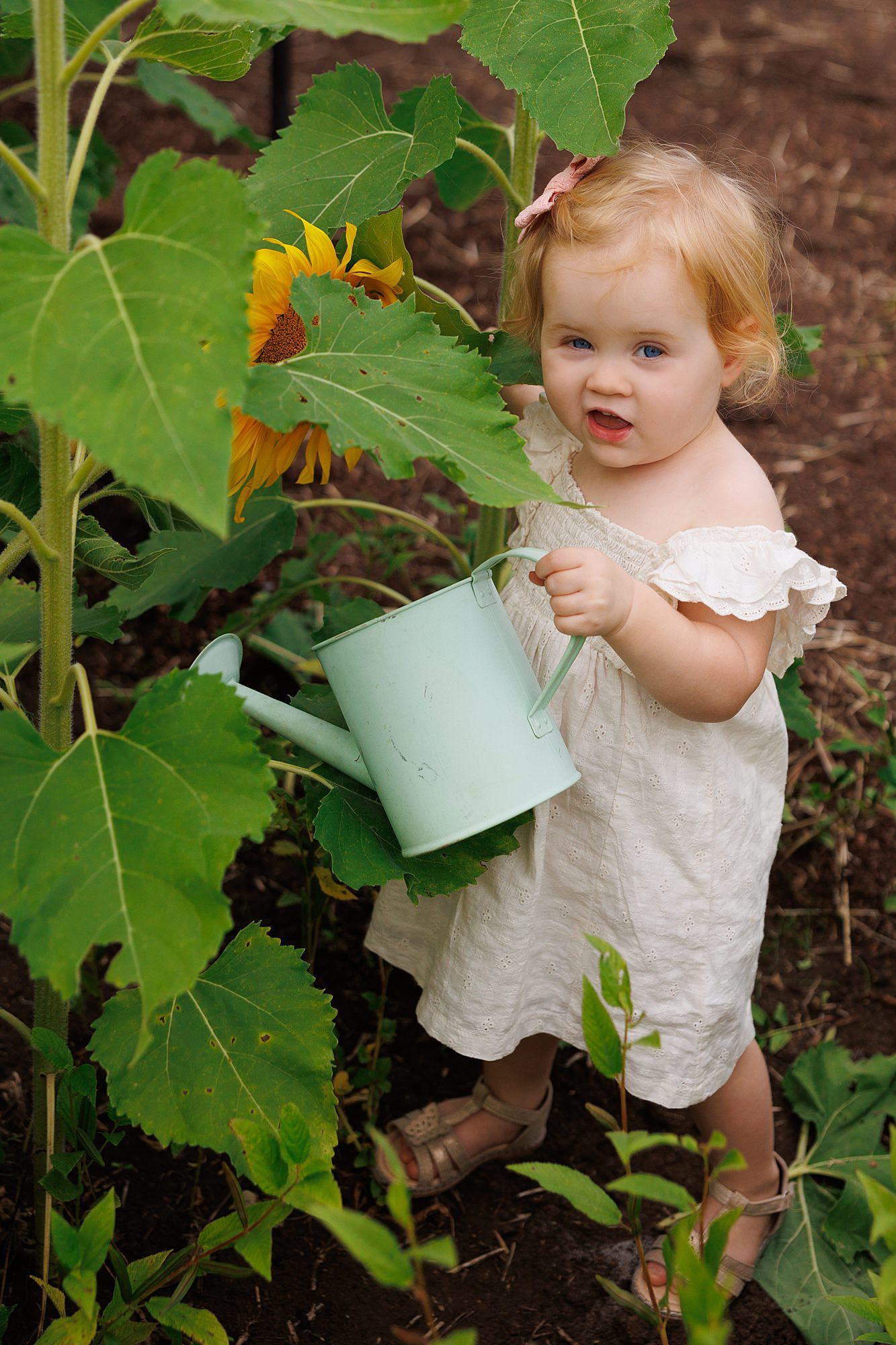A small gardener wearing a white dress holds a mint green watering can next to tall sunflower plants in a garden.