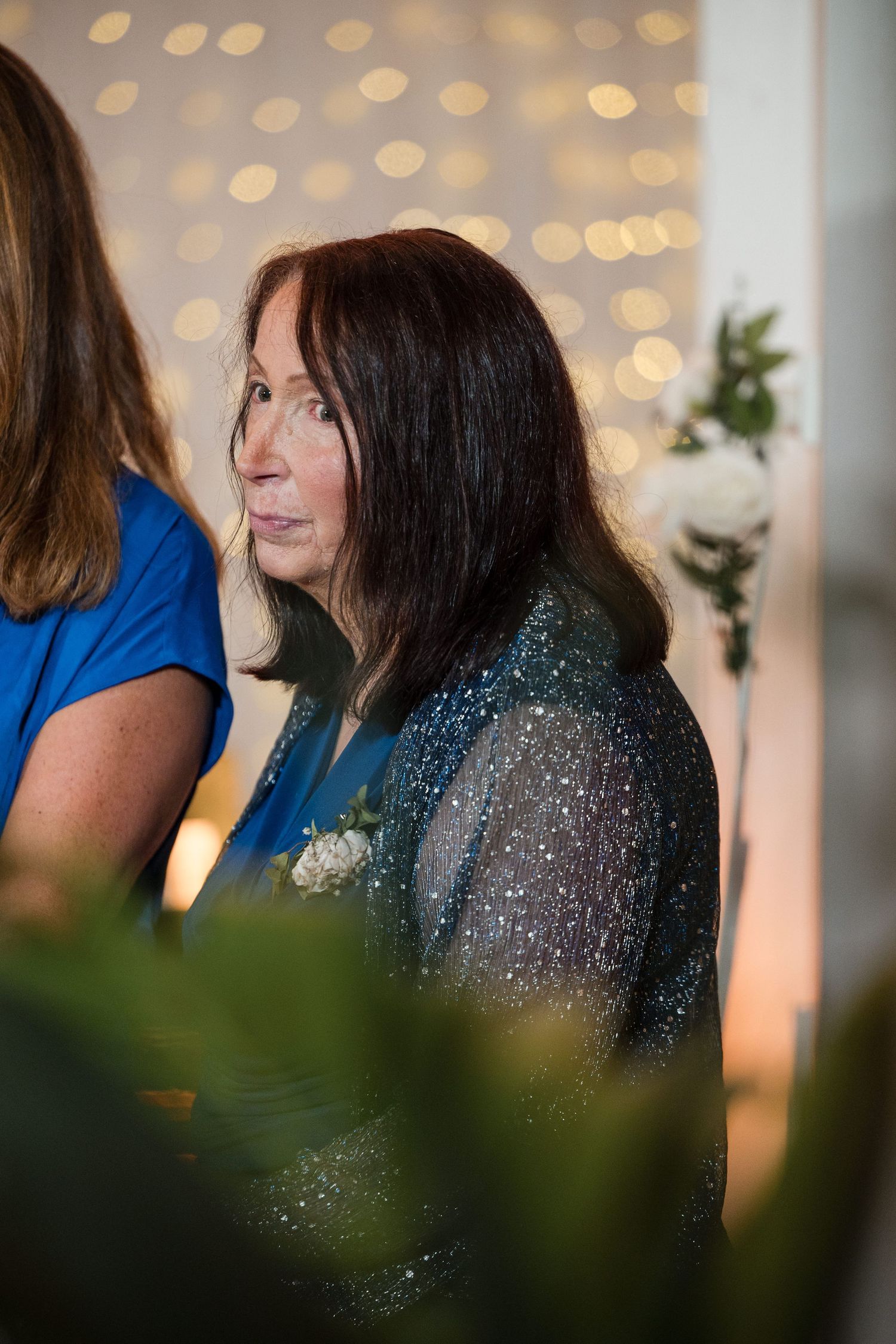Elegant older woman in a sparkly teal outfit with a corsage at a formal event with bokeh lights.