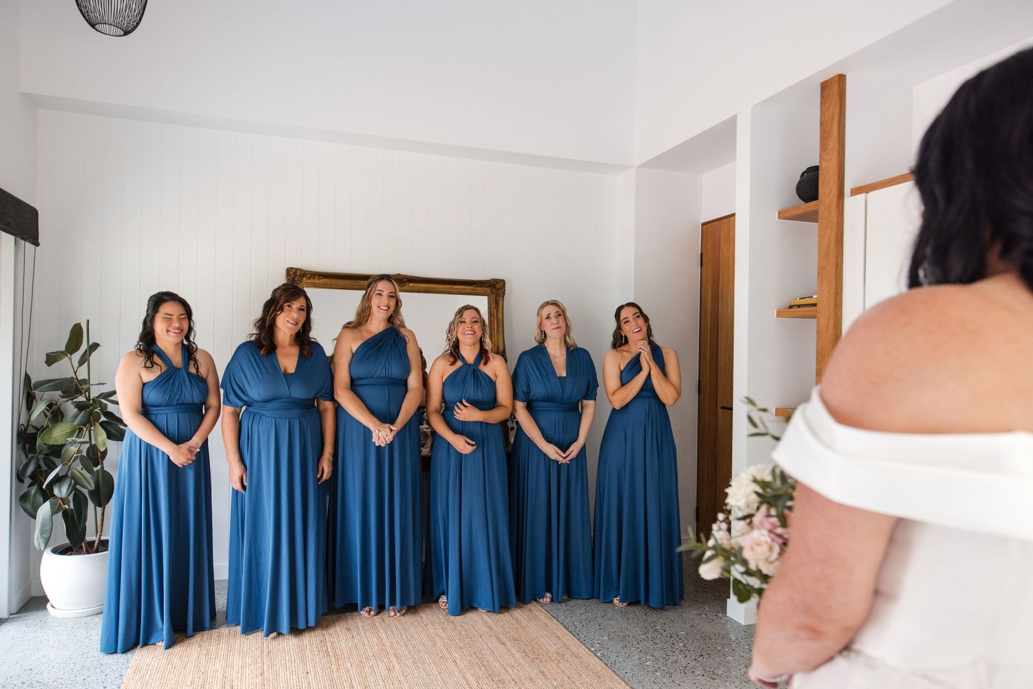 Six bridesmaids in blue gowns react emotionally as bride in white dress reveals her wedding look during bridal preparation.