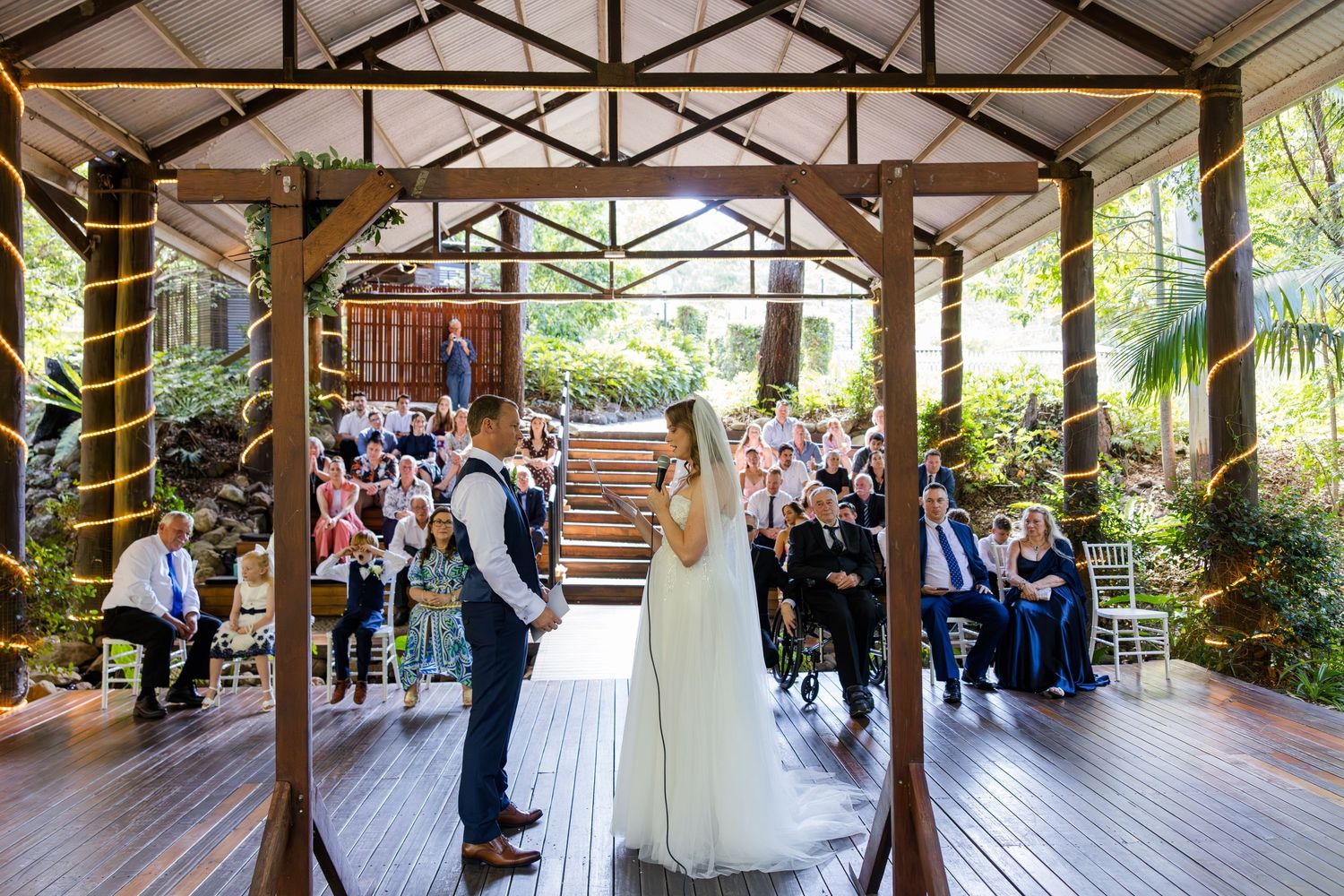 Wedding ceremony panorama in a rustic covered pavilion surrounded by lush greenery and wooden beams.