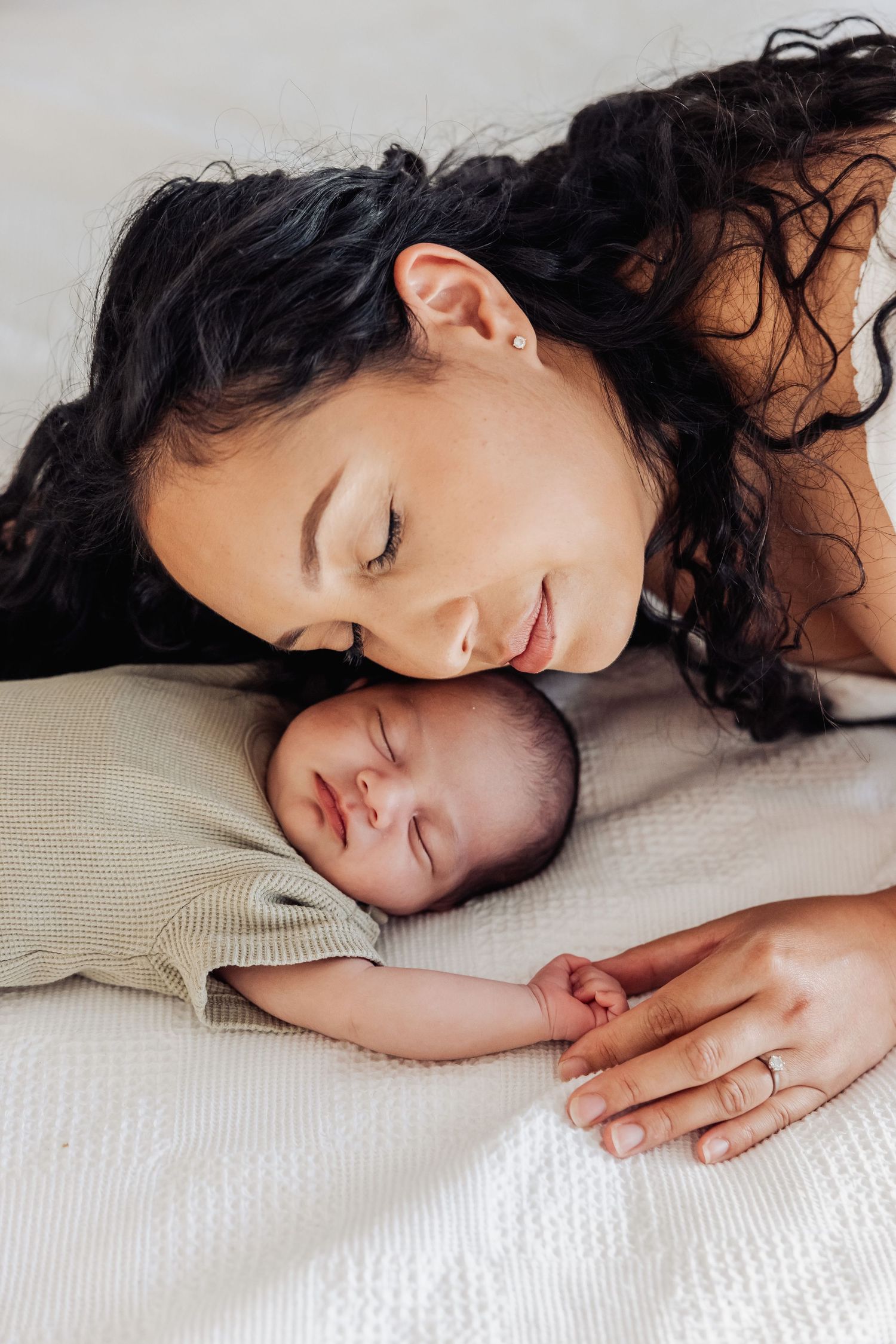 A mother with dark curly hair smiles tenderly at her sleeping newborn baby lying on a white blanket.