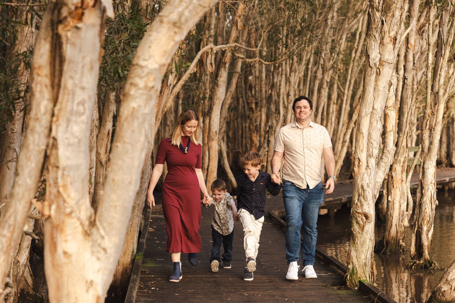 A family walks together holding hands along a wooden boardwalk surrounded by tall paperbark trees.