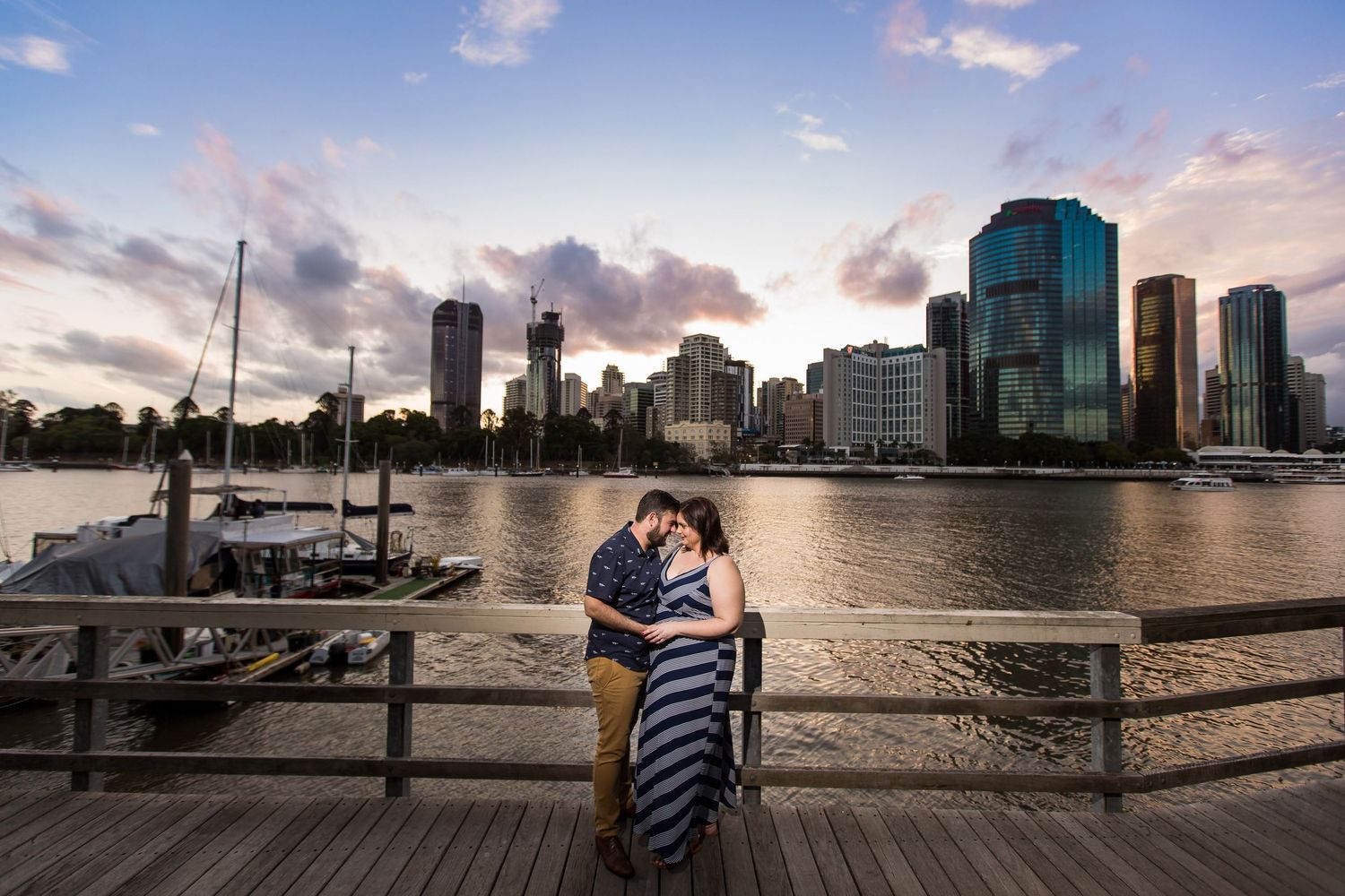 A couple stands on a waterfront boardwalk at sunset with Brisbane's modern skyline and river in the background.