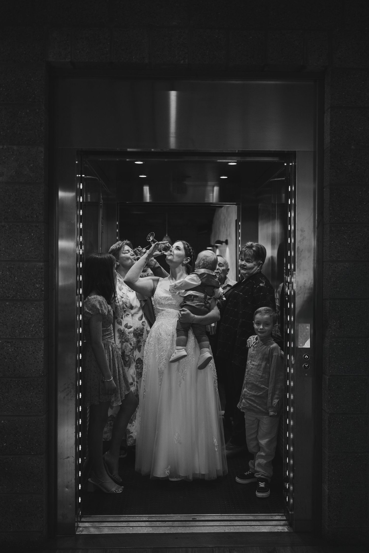 Black and white photo of a bride in a white gown surrounded by family and children inside a crowded elevator.