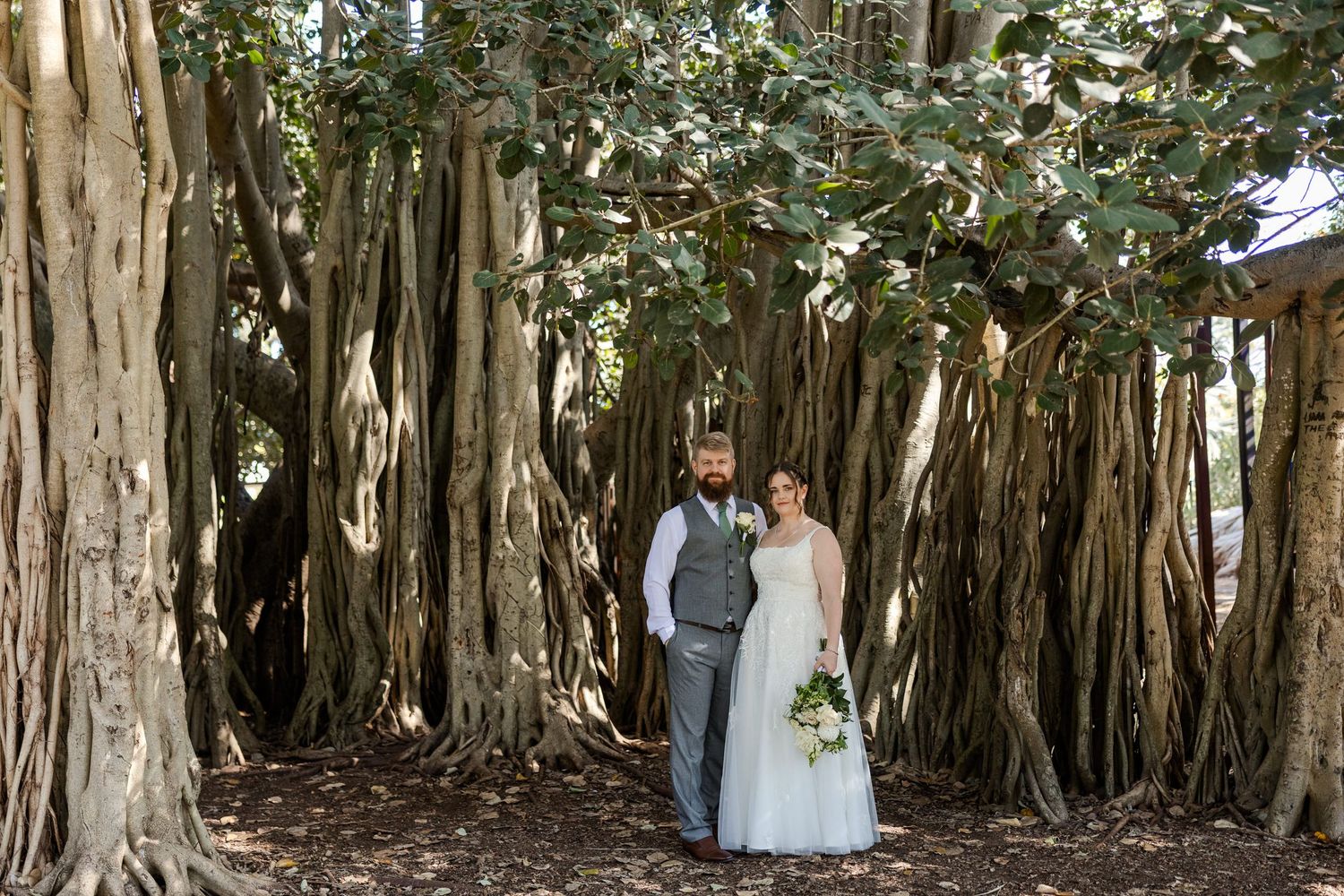 Bride and groom standing together under sprawling banyan tree roots at outdoor wedding ceremony.