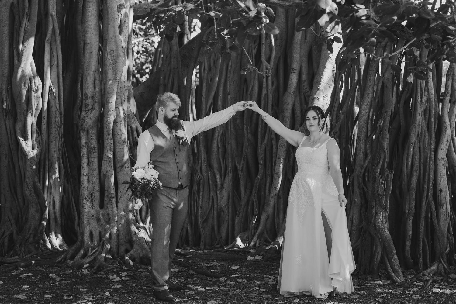 Black and white photo of bride and groom holding hands under a large banyan tree during outdoor wedding ceremony.