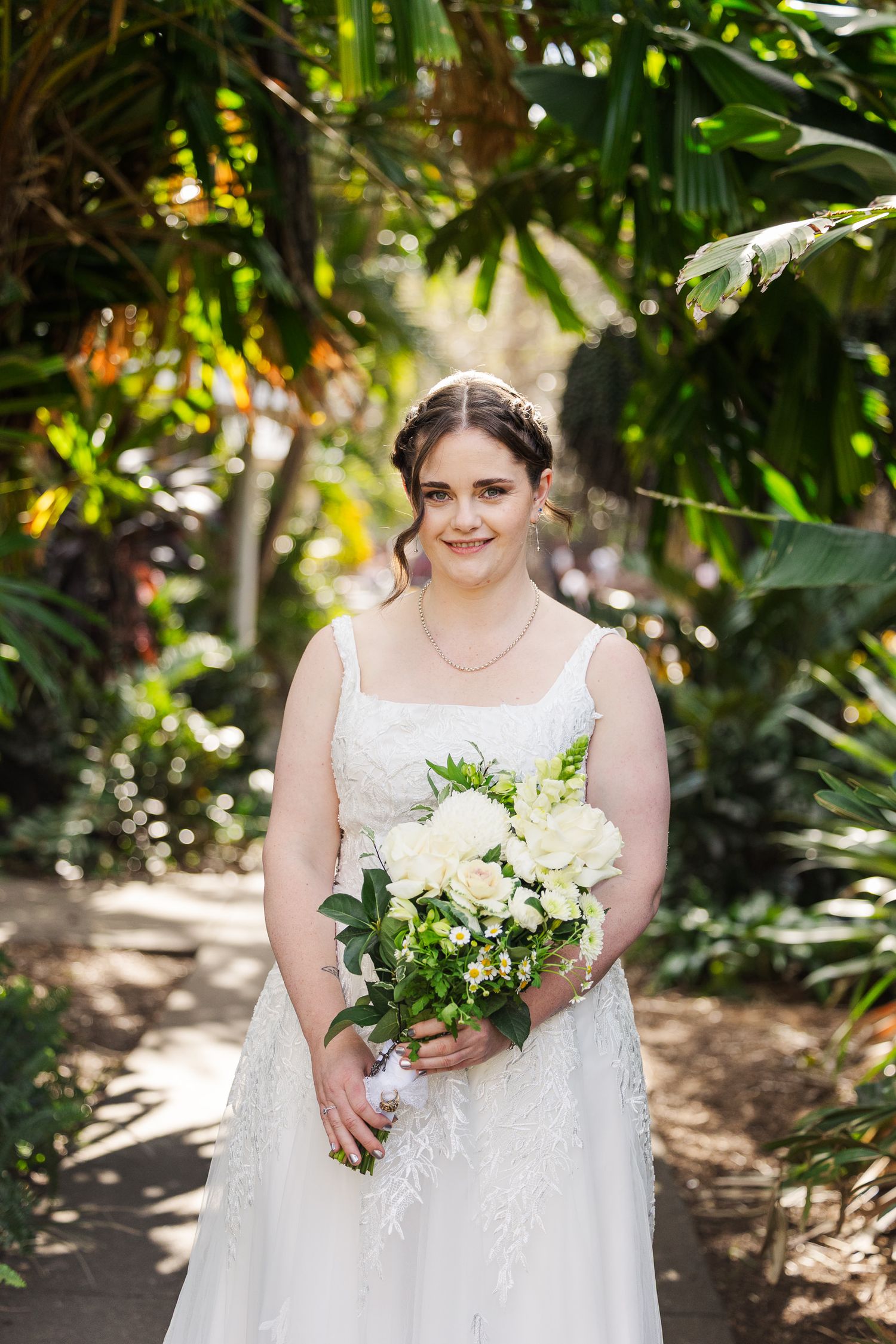 Bride in white lace gown holding white bouquet smiles in lush tropical garden setting.