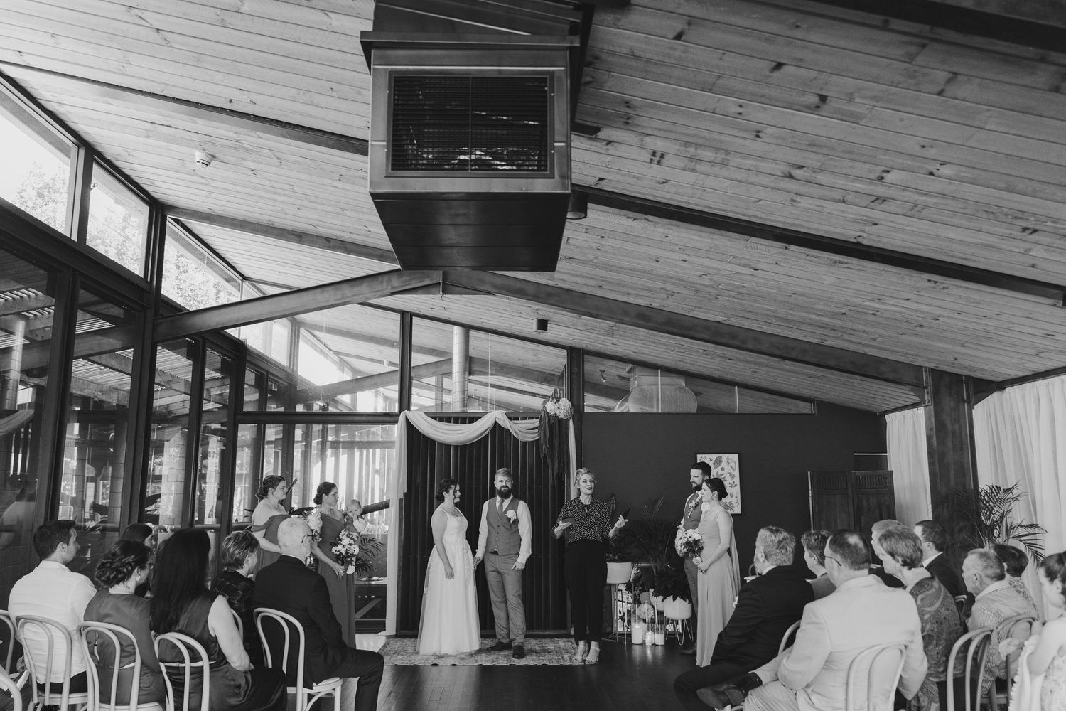 Black and white photo of an elegant indoor wedding ceremony with guests seated in a rustic wood-beamed venue.
