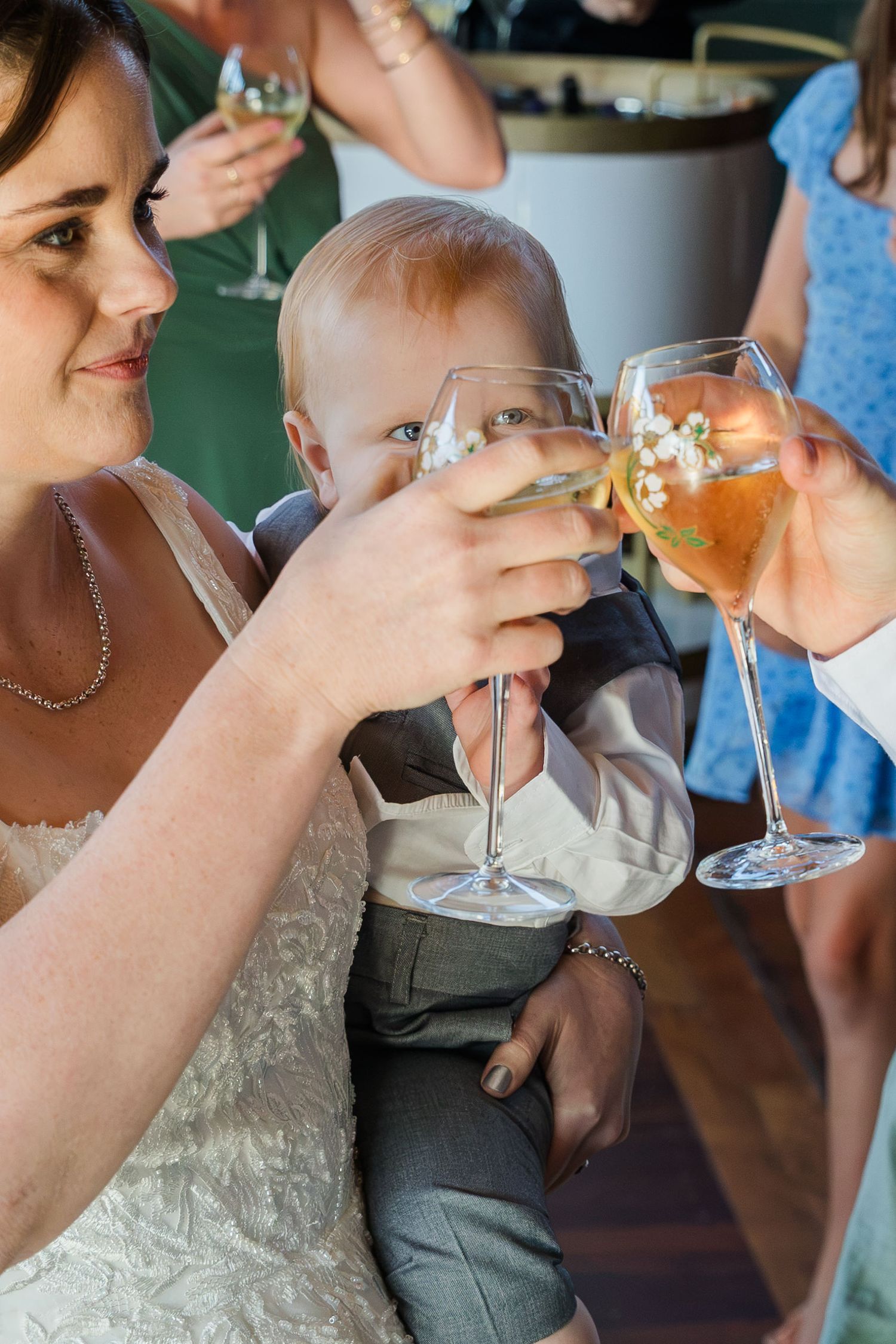 Bride holding baby during wedding toast, clinking wine glasses with guests at a celebratory reception.
