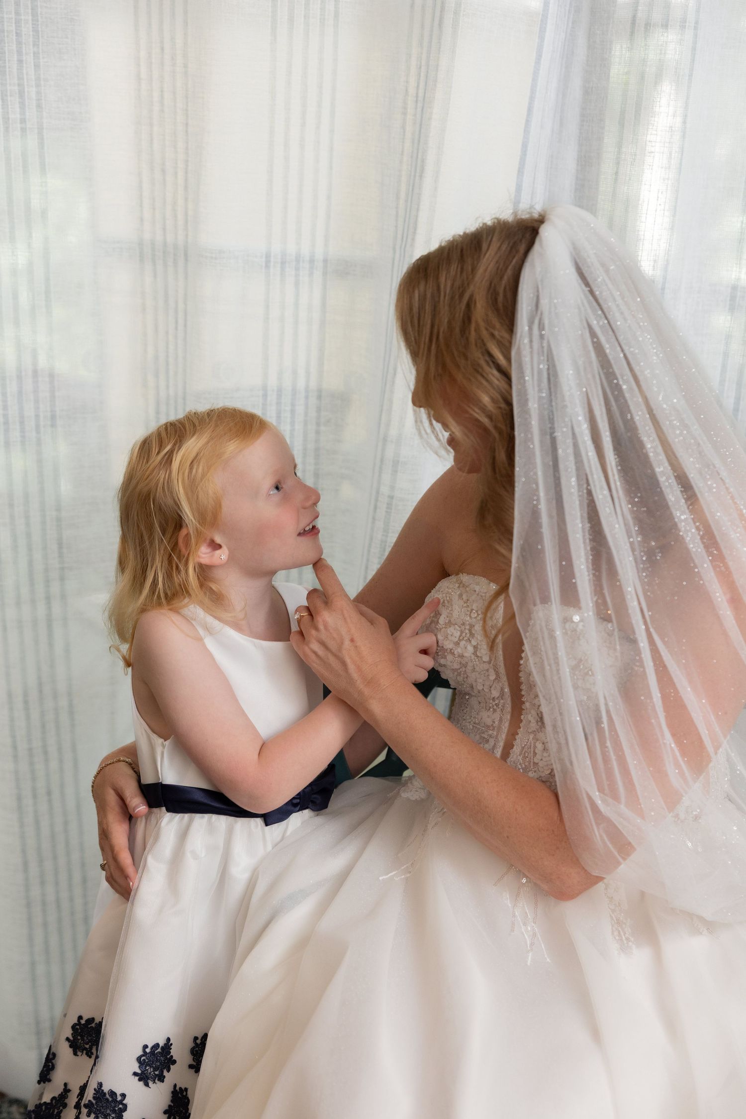 A bride in a white gown and veil shares an intimate moment with a young child wearing a white dress with navy trim.