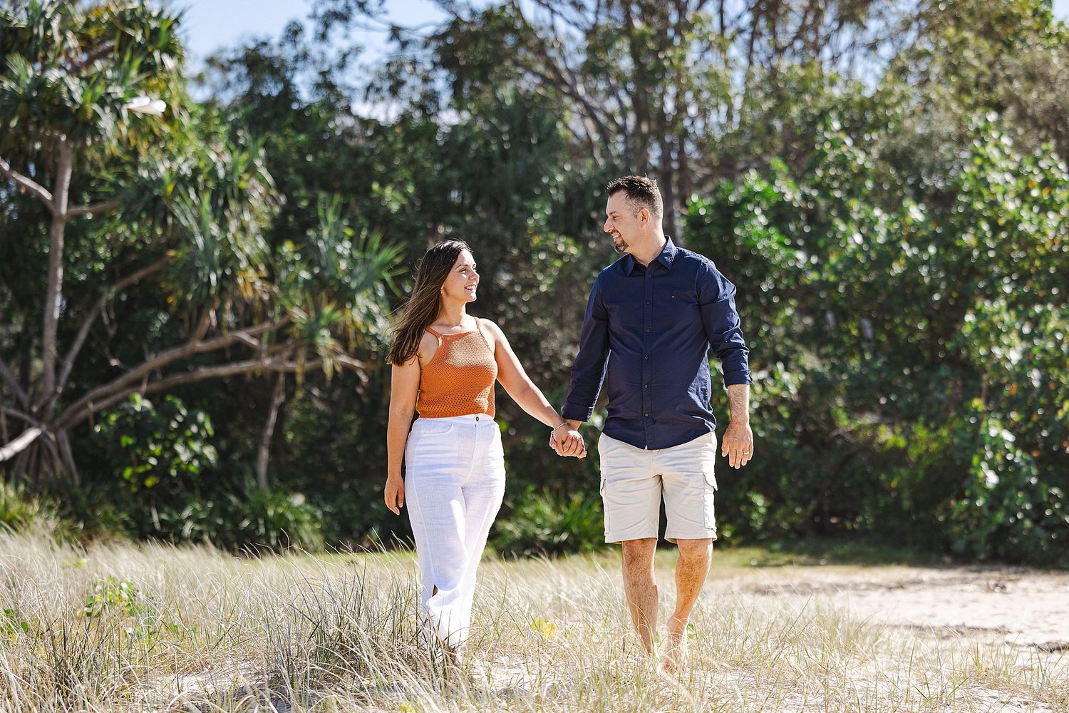 Couple walking hand in hand on a dirt path through a scenic wooded area surrounded by eucalyptus trees on a sunny day.