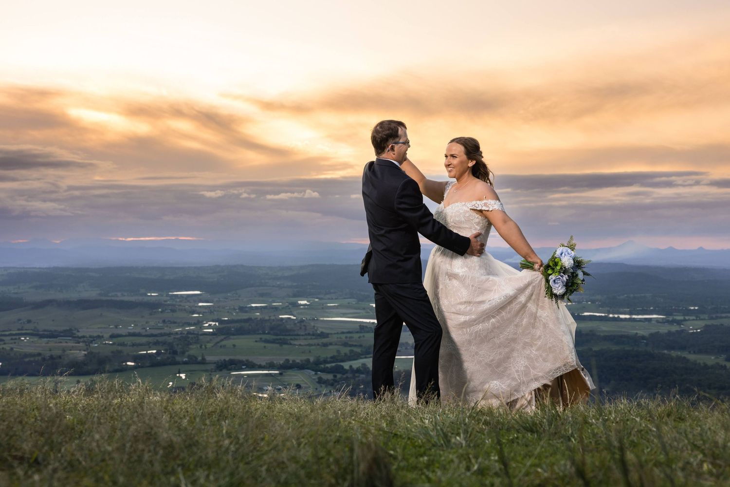 A couple stands together on a hilltop during sunset with a scenic mountain landscape in the background.