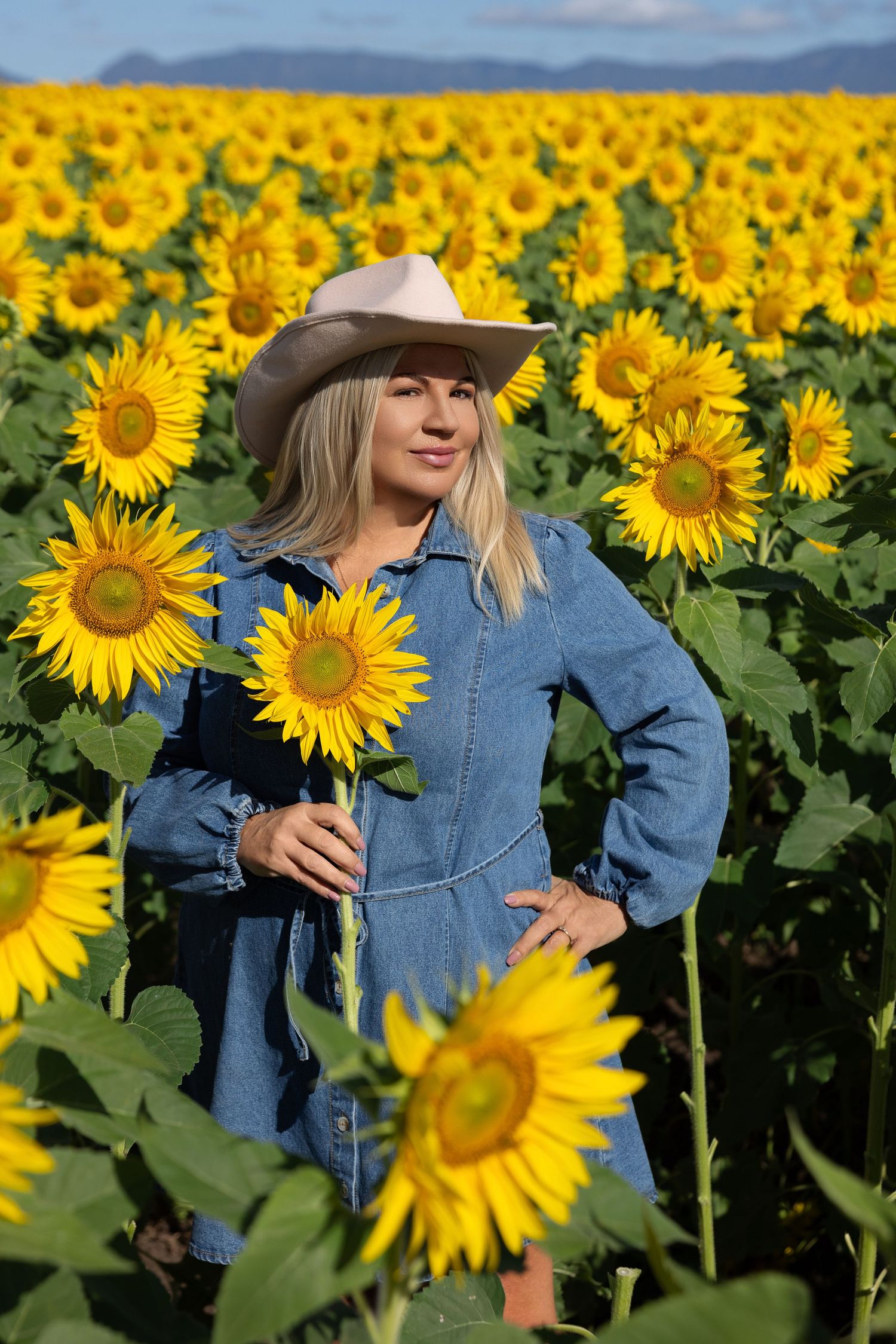 Blonde woman in denim dress and cowboy hat holding a sunflower in a vibrant sunflower field.