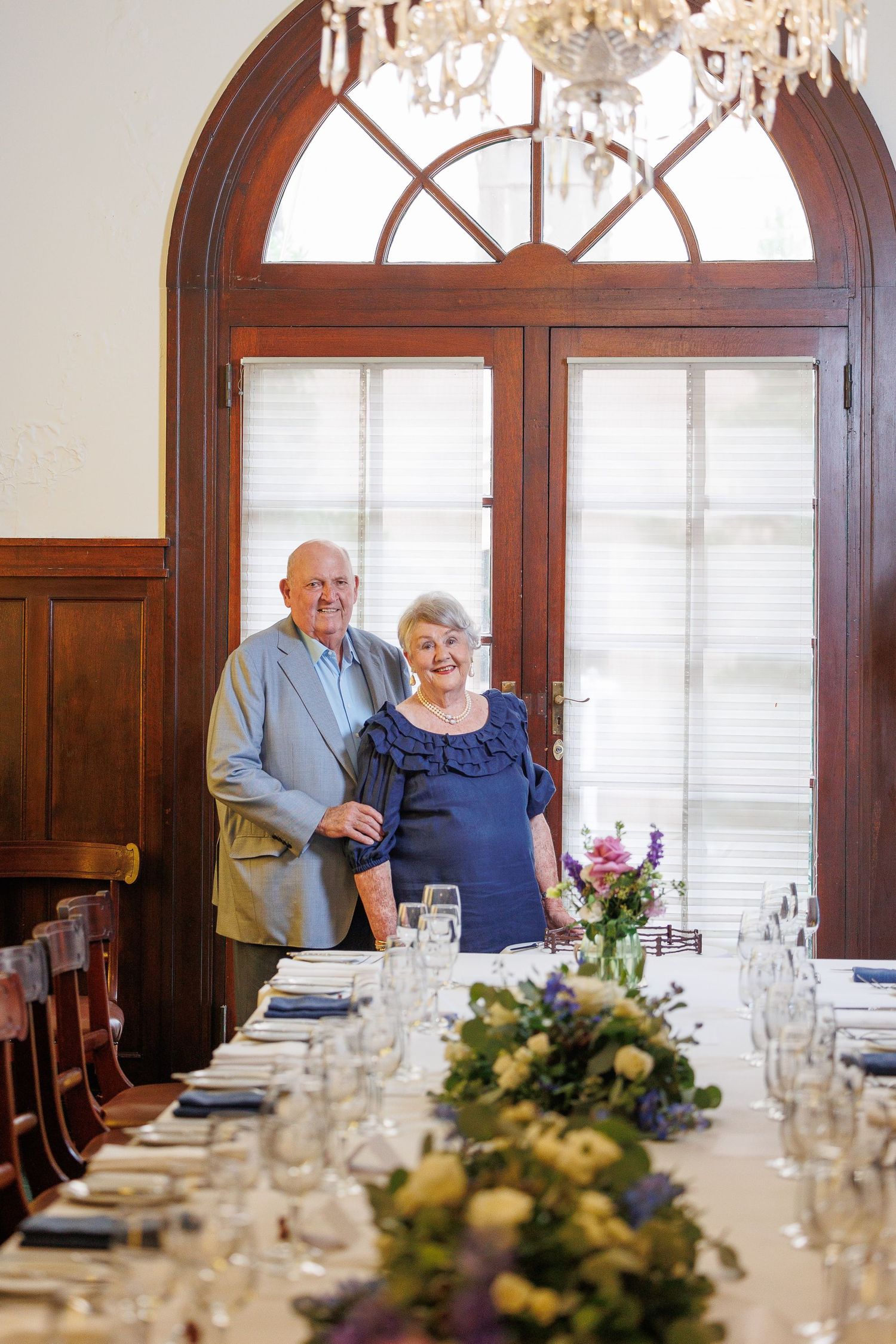 An elegant dining room with crystal chandelier features a long table decorated with floral centerpieces and formal place settings.
