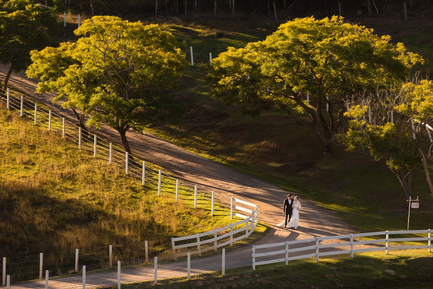 Couple walking along scenic hillside path with white fence and golden trees.