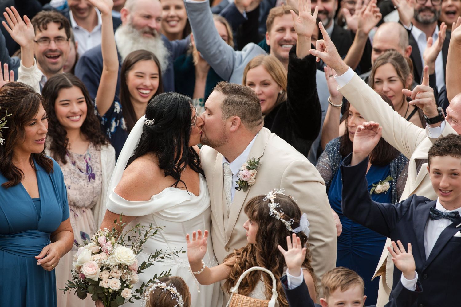 Bride and groom share a kiss surrounded by cheering wedding guests, with flower girl nearby.