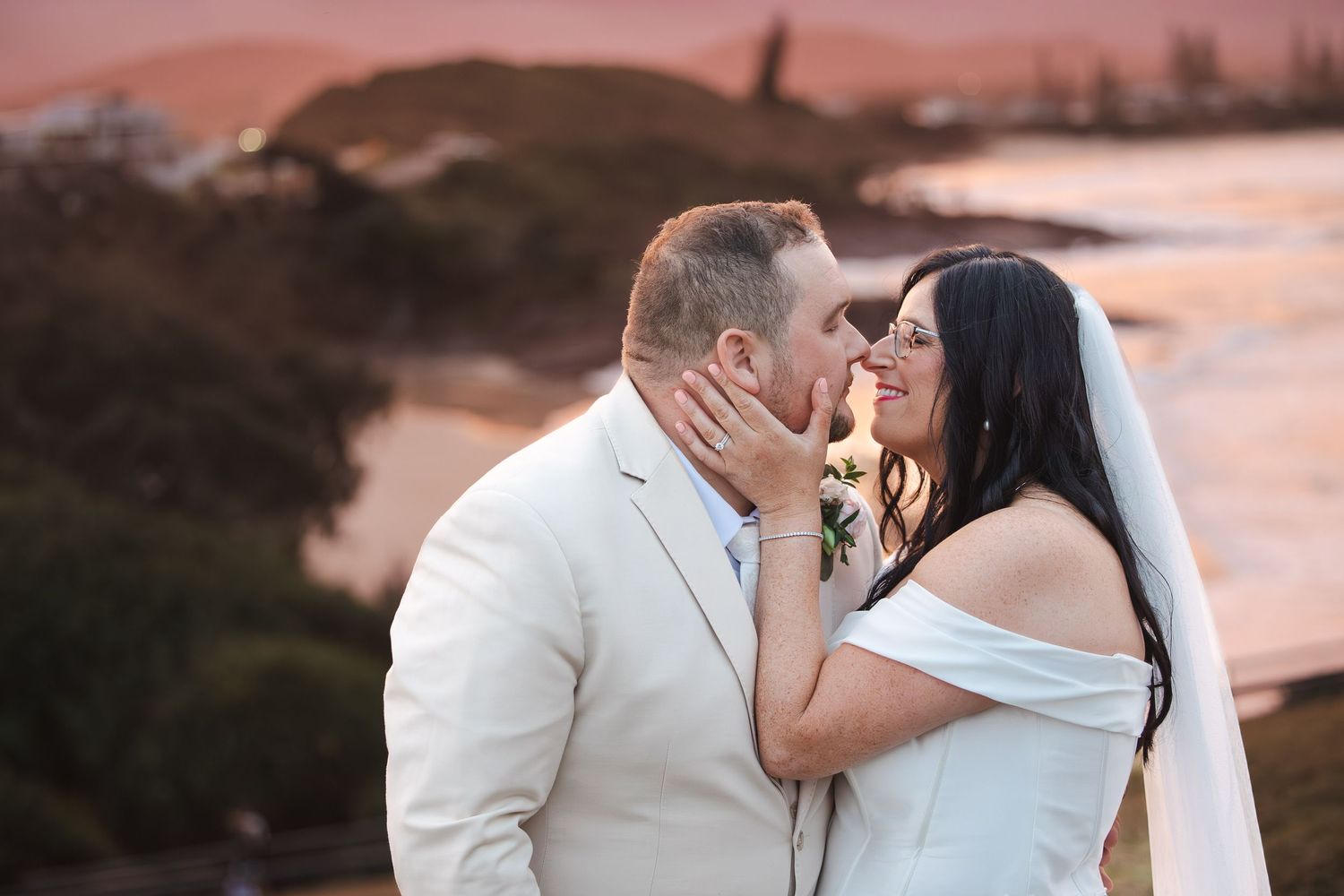 Bride and groom share a romantic kiss at sunset with ocean views in background at beach wedding.