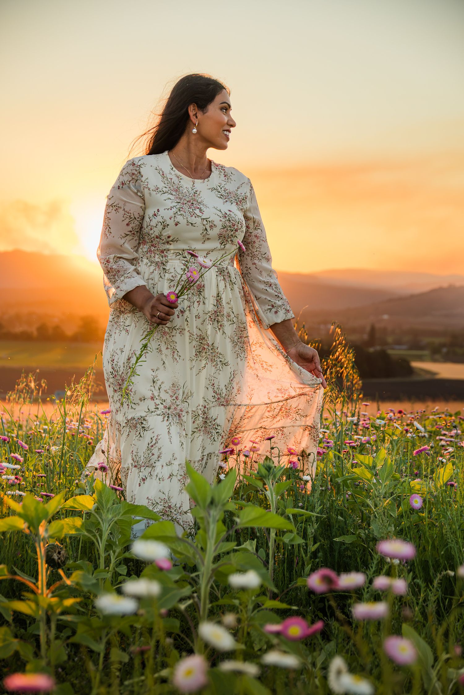 Woman in floral white dress standing in wildflower field at golden sunset with mountain backdrop.