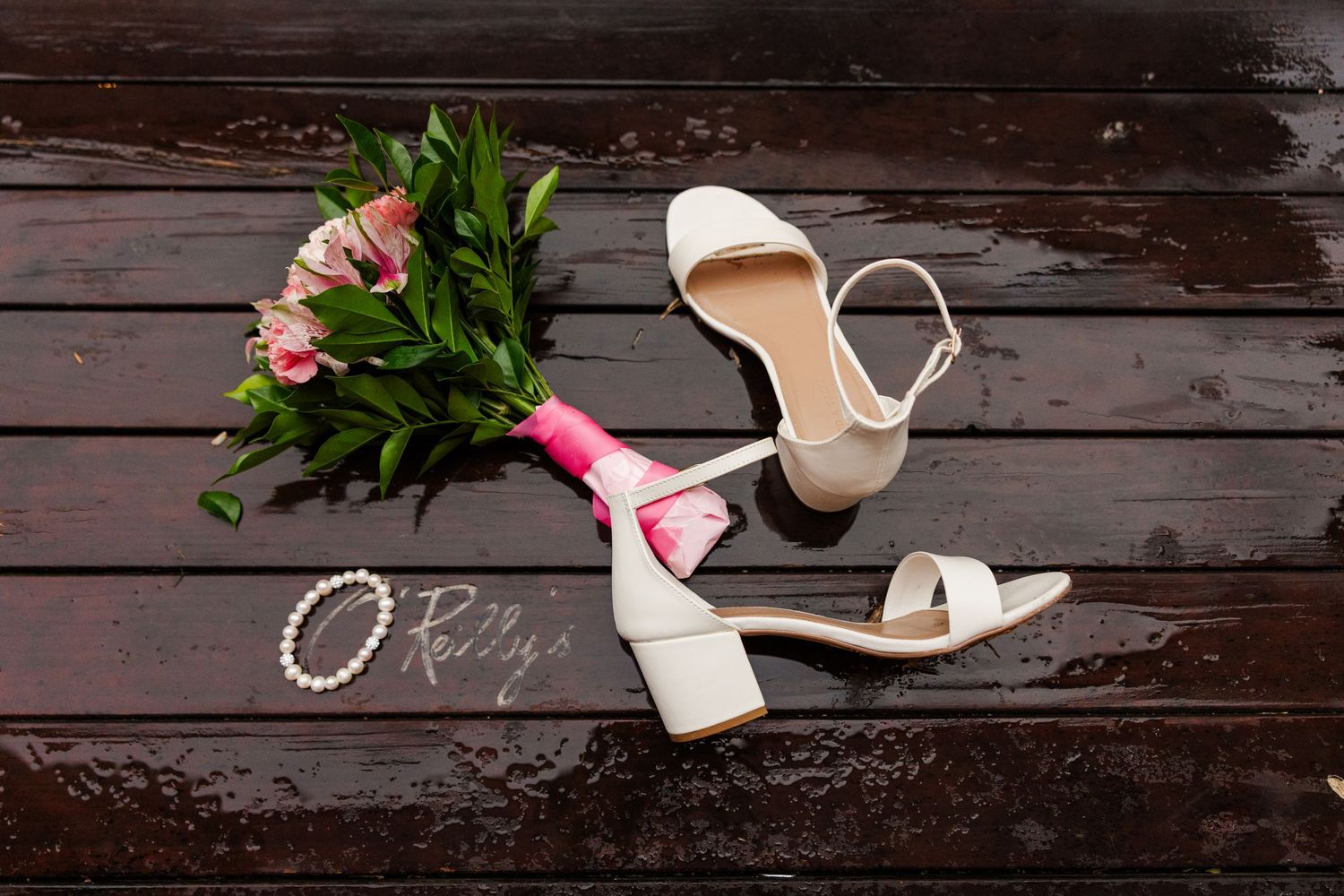 White bridal heels beside a pink flower bouquet and pearl bracelet on a dark wooden surface.