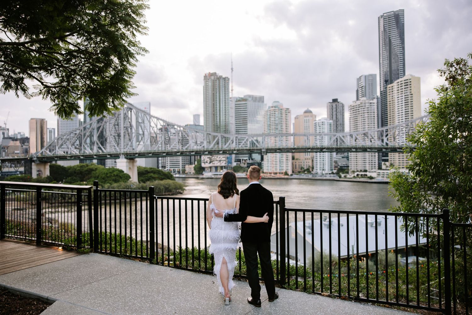 People stroll along a scenic waterfront promenade with city skyline views and bridge in the background.