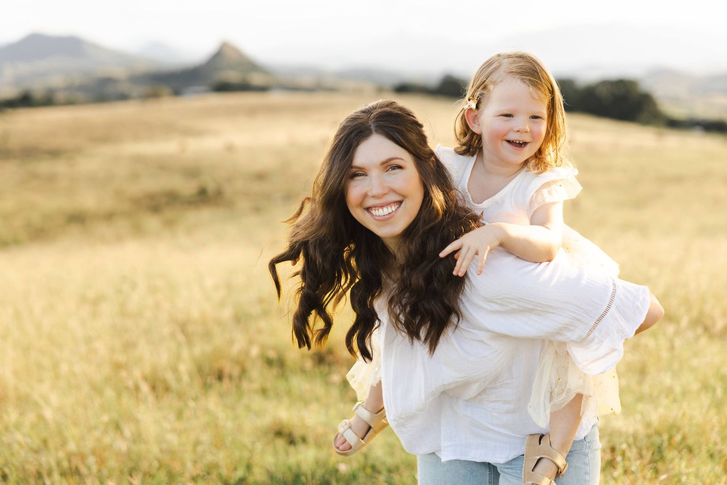 A pair enjoying a playful piggyback ride in a sunlit golden field of tall grass with mountains in the distance.