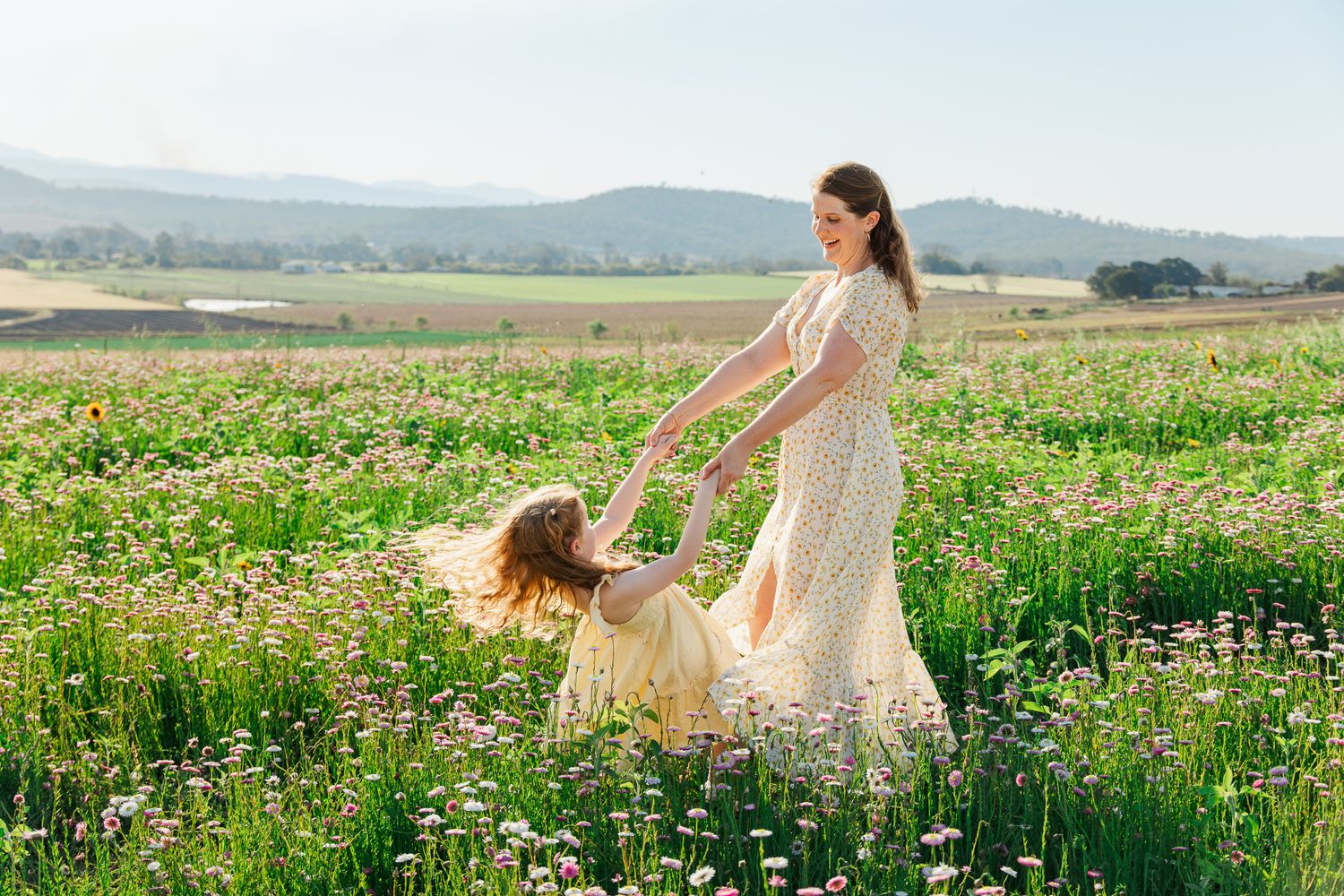Mother and daughter in floral dresses twirling in a sunny wildflower meadow with mountains in the background.