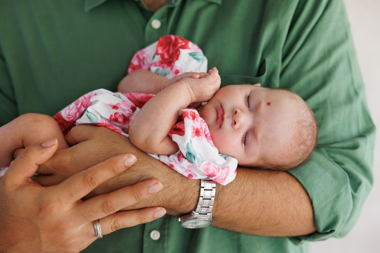 A tiny newborn baby in a floral onesie rests peacefully in an adult's arms wearing a green shirt and watch.