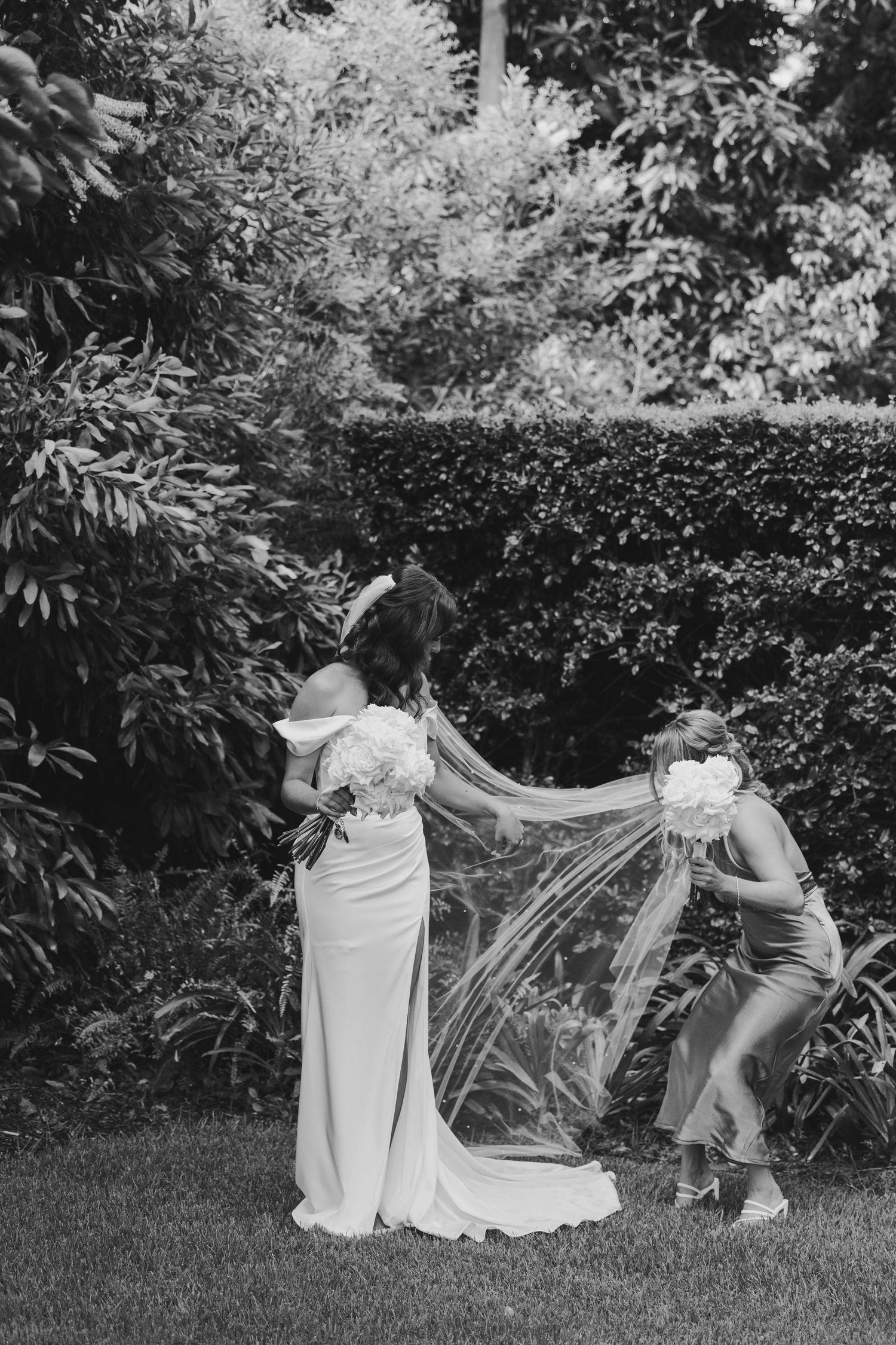 A black and white photograph showing a woman in a long white wedding dress with a flowing veil in a garden setting.