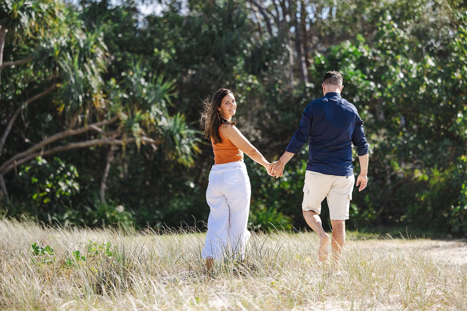 A couple holding hands as they walk along a grassy outdoor path near trees on a sunny day.