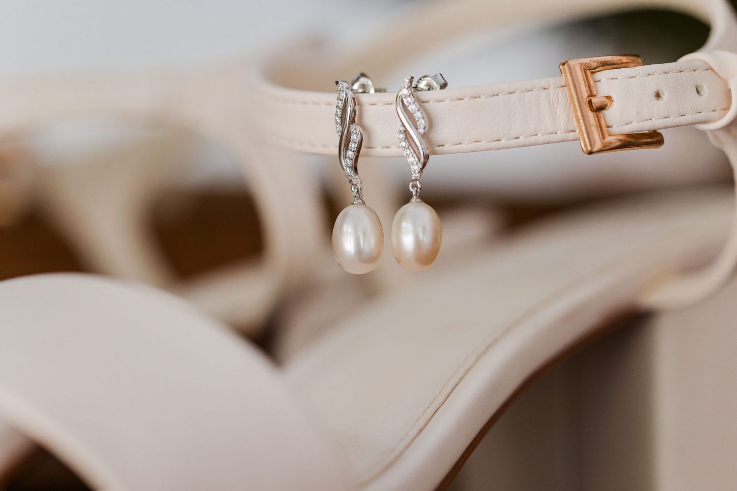 Pearl drop earrings on silver hooks displayed on beige strappy heeled sandals with gold buckle detail.