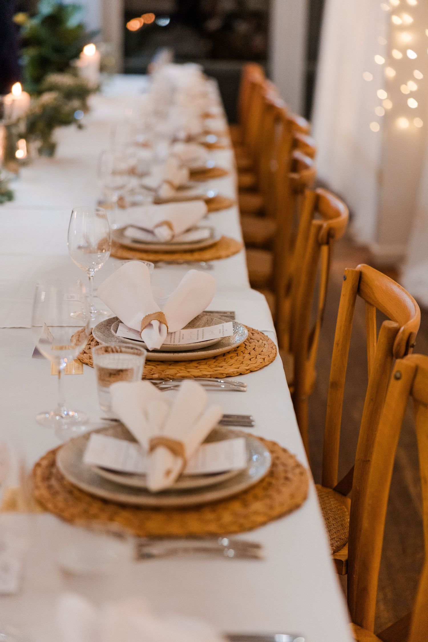 Elegant long dining table with woven placemats, white napkins, glassware, and warm candlelight for a formal event.