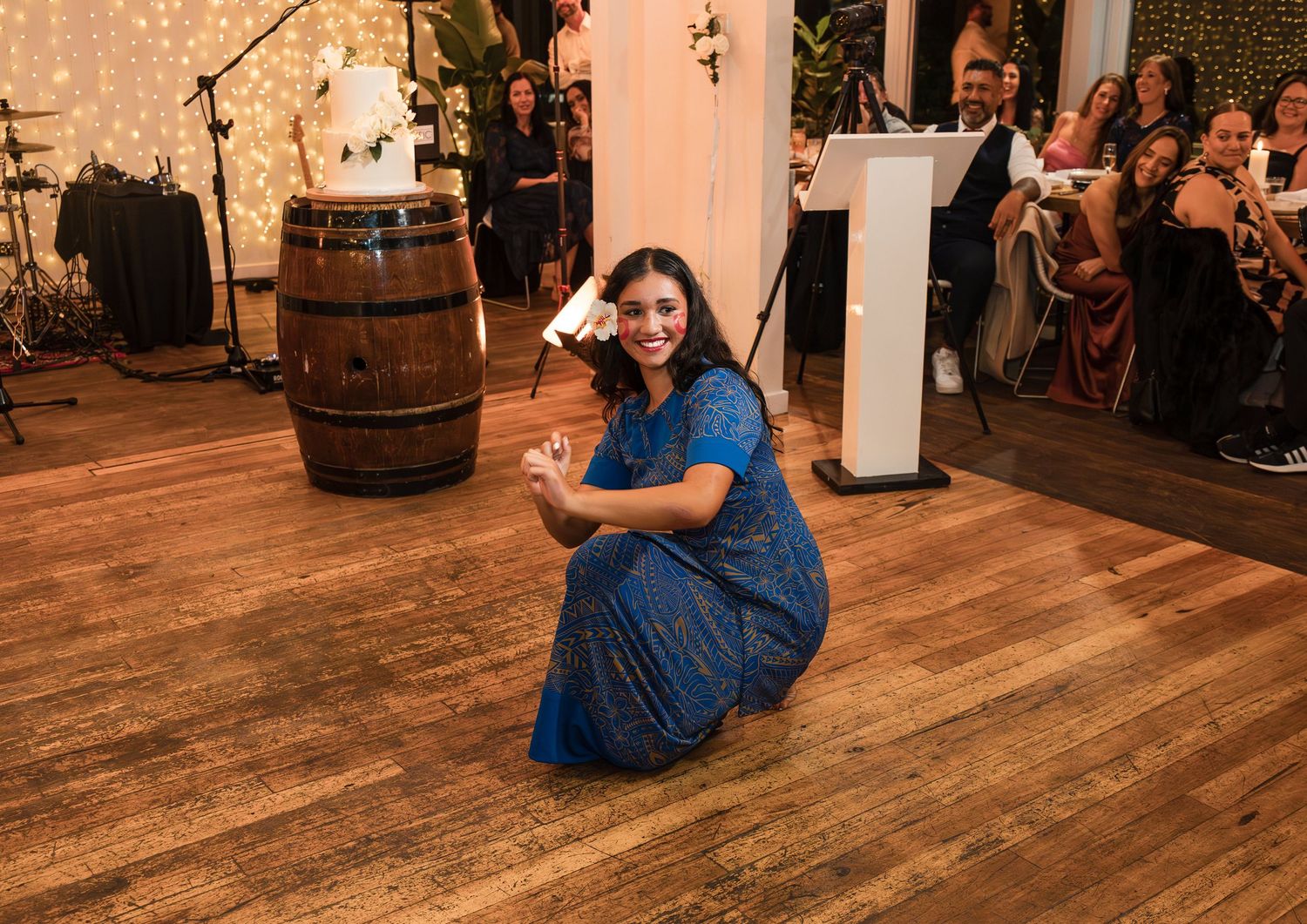Woman in blue saree crouching on dance floor at indoor wedding reception with barrel and wedding cake in background.