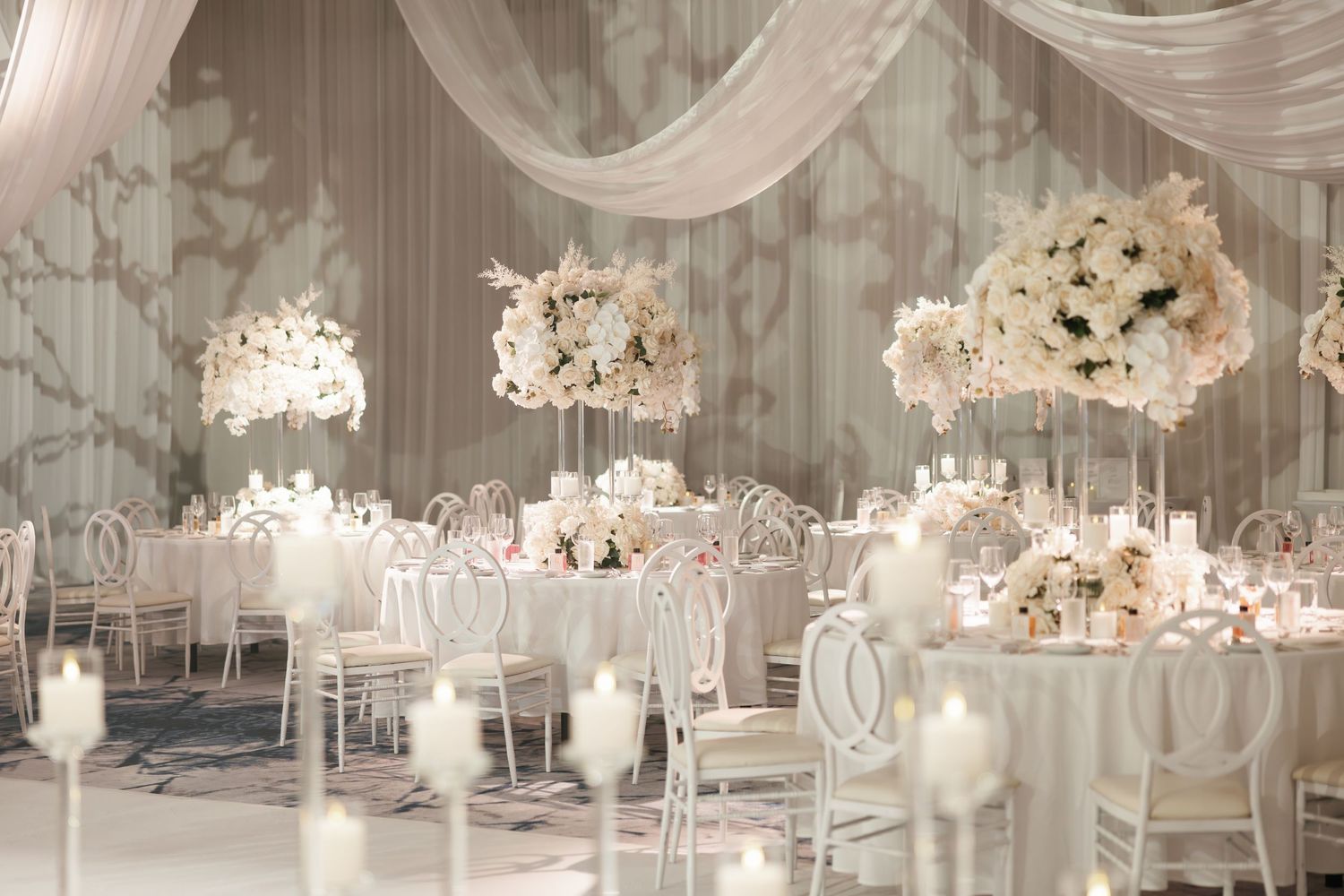 Elegant white wedding reception hall with crystal chandeliers, draped fabric, and floral centerpieces on round tables.