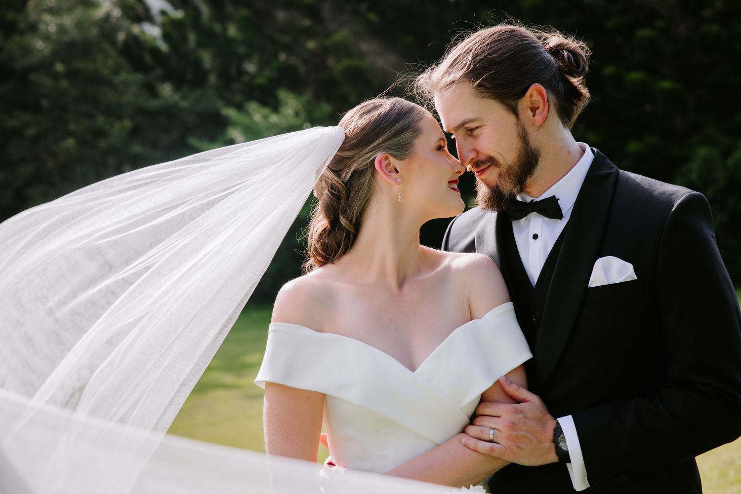 A romantic wedding couple shares an intimate moment in a garden setting with a flowing white veil in the breeze.