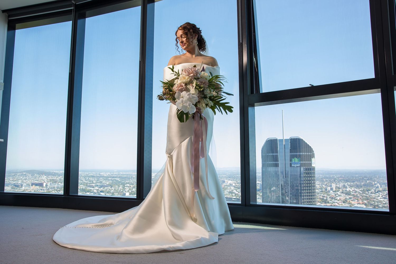 A bride in a flowing white wedding gown stands by large windows overlooking a cityscape on her special day.
