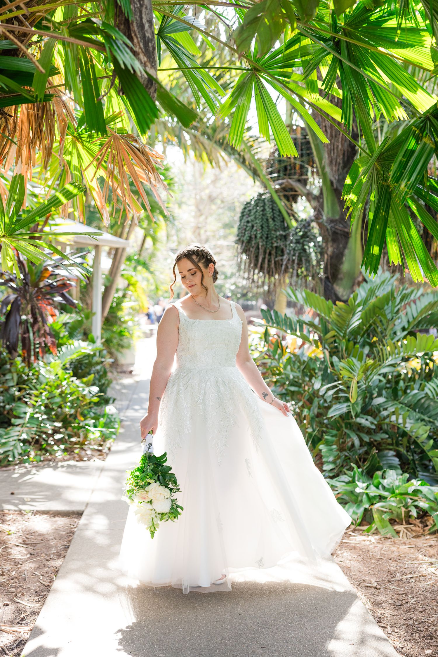Bride in white lace ball gown holding bouquet walks along tropical garden path surrounded by lush green palms.