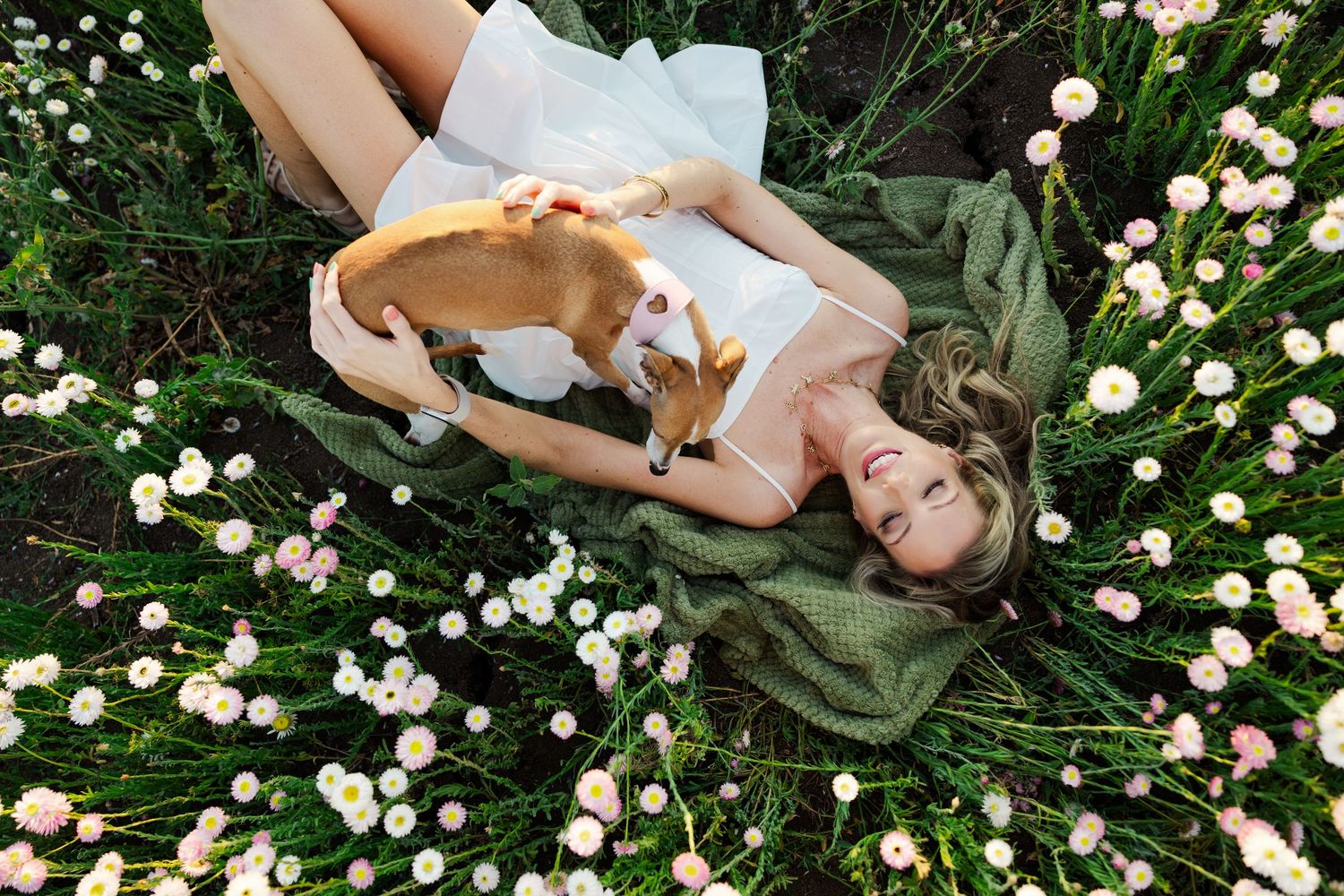 A smiling woman in a white dress lies in a flower field cuddling a small dog among white ranunculus blooms.