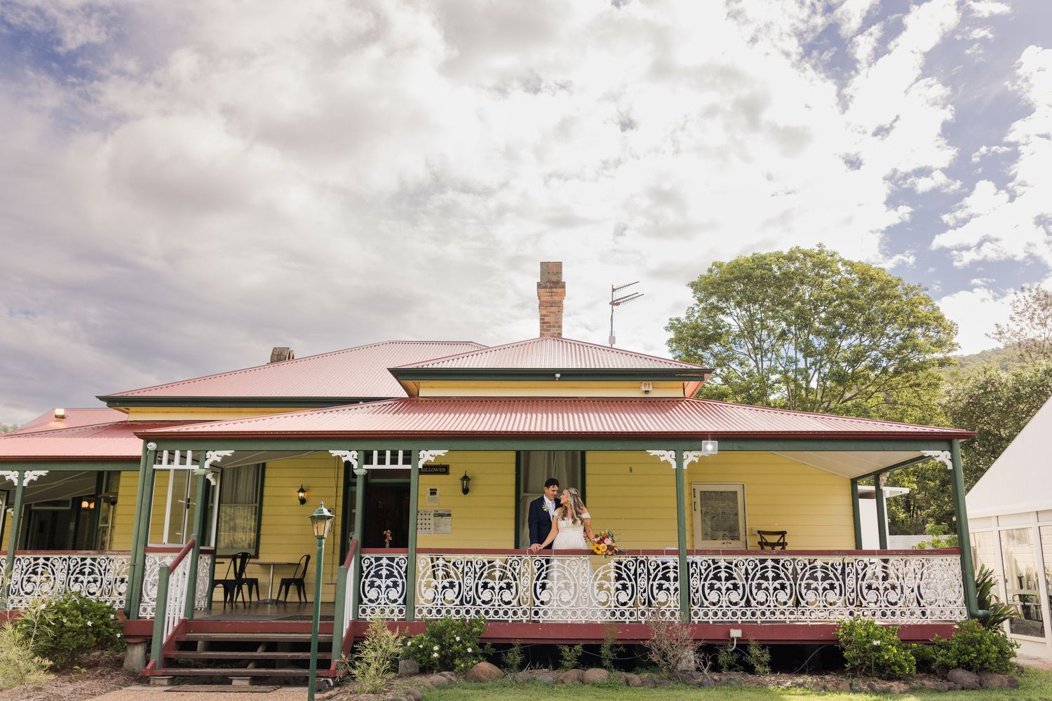Couple embracing on the veranda of a charming yellow Victorian heritage home with decorative ironwork trim.