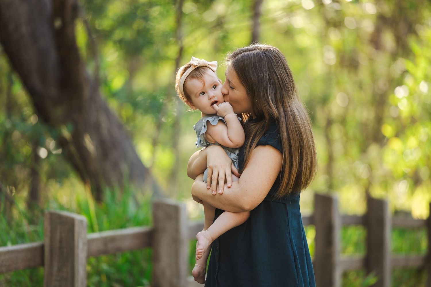 A mother kisses her baby boy outdoors on a wooden bridge surrounded by lush green trees.